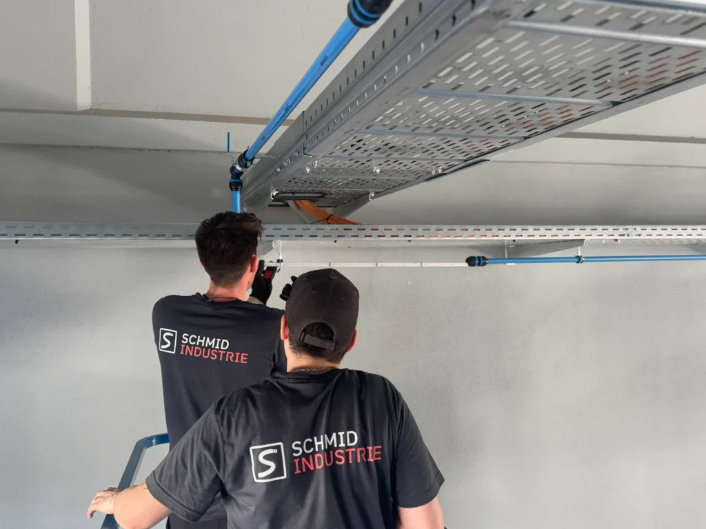 zwei Personen vor einer blauen Aluminium Druckluftleitung mit einem Schmid Industrie T-Shirt vermessen ein stück Rohrleitung mit dem Meterstab. Die Bauteile aus der Drucklufttechnik sind an einer Elektrotrasse montiert.