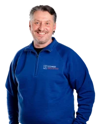 Ein Mann mit grauen Haaren lächelt und hat einen Blauen Pullover an mit einem Schmid Industrie Logo auf seiner linken Brust. Er hat graue Haare und Trägt einen Ohrring und lächelt.
